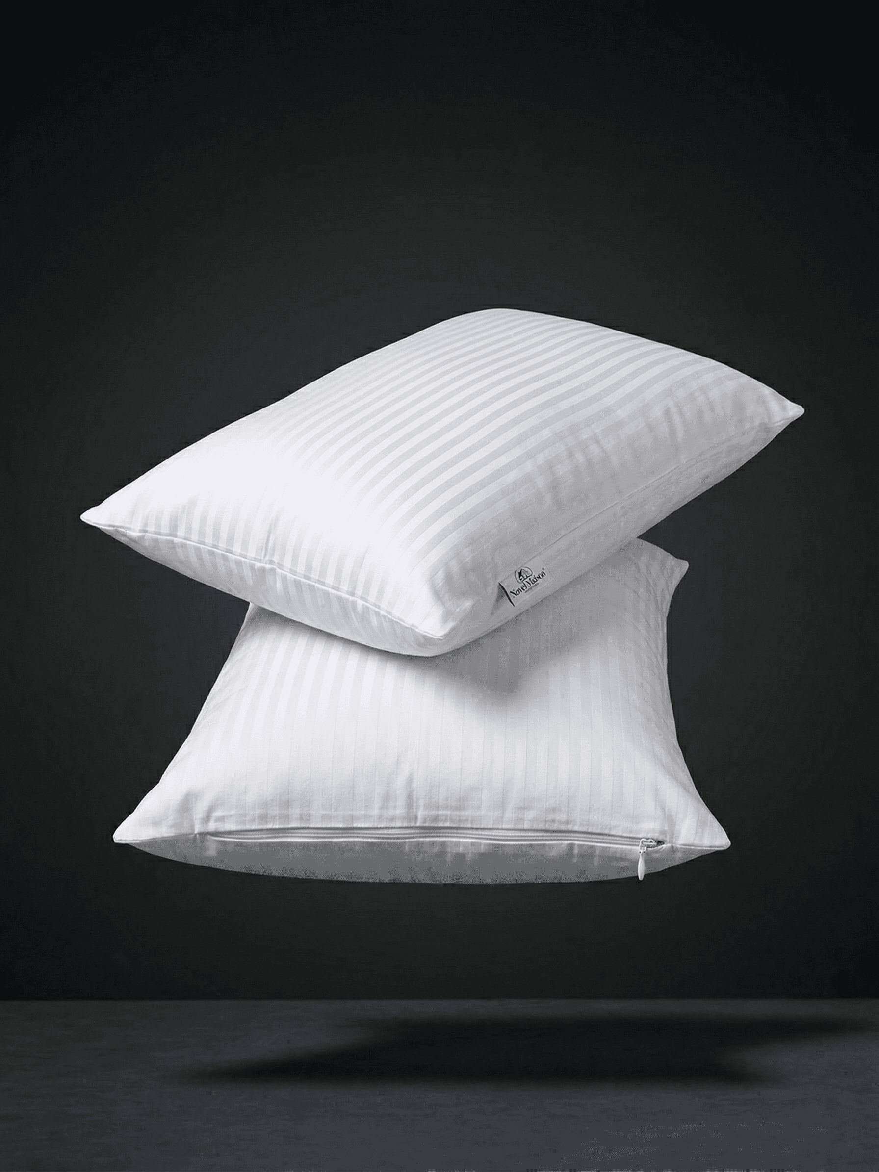 Una fotografia professionale di alta qualità e pulita di due federe di cotone bianco, impilate in modo minimalista e che fluttuano in un ambiente di studio scuro con illuminazione drammatica. Le federe sono di un bianco pulito, ben stirate e la loro forma è ben definita, mostrando la consistenza del tessuto in cotone di qualità. Una leggera ombra morbida è proiettata sulla superficie grigia sottostante, creando un effetto di profondità e leggerezza. Lo sfondo è un nero profondo e sfumato. Questa immagine es