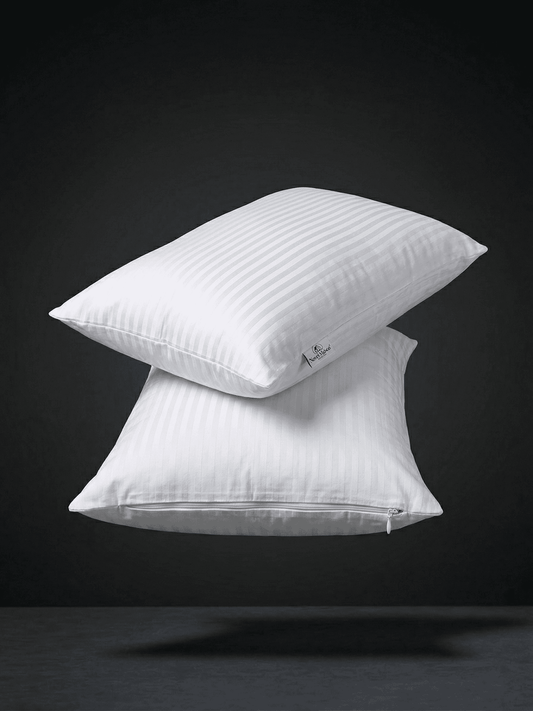 Una fotografia professionale di alta qualità e pulita di due federe di cotone bianco, impilate in modo minimalista e che fluttuano in un ambiente di studio scuro con illuminazione drammatica. Le federe sono di un bianco pulito, ben stirate e la loro forma è ben definita, mostrando la consistenza del tessuto in cotone di qualità. Una leggera ombra morbida è proiettata sulla superficie grigia sottostante, creando un effetto di profondità e leggerezza. Lo sfondo è un nero profondo e sfumato. Questa immagine es