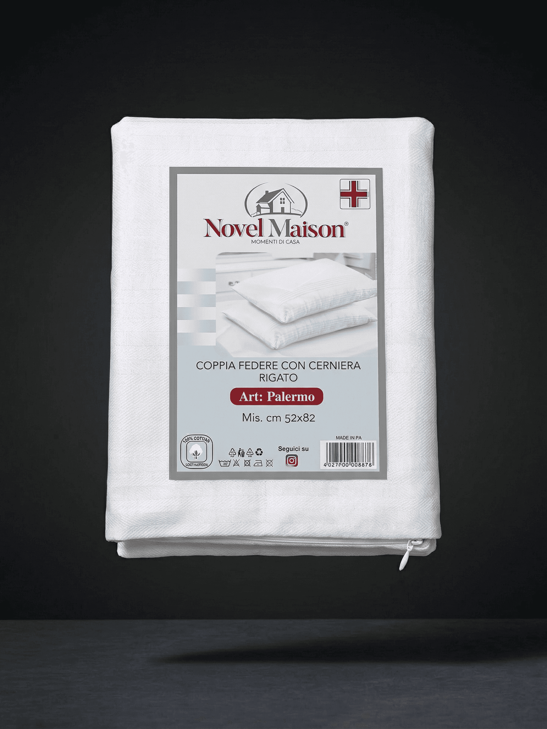 In primo piano, una fotografia di prodotto professionale e nitida del packaging di una federa per cuscino in cotone Novel Maison "copri cuscino", piegata e sigillata in un imballaggio di plastica e cartone. L'imballaggio in cartone mostra chiaramente il logo "NOVEL MAISON" con un'icona di una piccola casa e un dettaglio della bandiera italiana (o indicazione di "Made in Italy"). Il testo italiano sull'imballaggio dice "COPRI CUSCINO Con cerniera In cotone Mis. cm 50x80 1 federa". Sull'imballaggio c'è anche 
