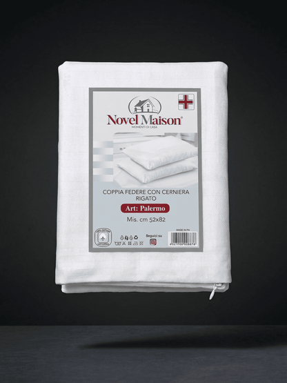 In primo piano, una fotografia di prodotto professionale e nitida del packaging di una federa per cuscino in cotone Novel Maison "copri cuscino", piegata e sigillata in un imballaggio di plastica e cartone. L'imballaggio in cartone mostra chiaramente il logo "NOVEL MAISON" con un'icona di una piccola casa e un dettaglio della bandiera italiana (o indicazione di "Made in Italy"). Il testo italiano sull'imballaggio dice "COPRI CUSCINO Con cerniera In cotone Mis. cm 50x80 1 federa". Sull'imballaggio c'è anche 