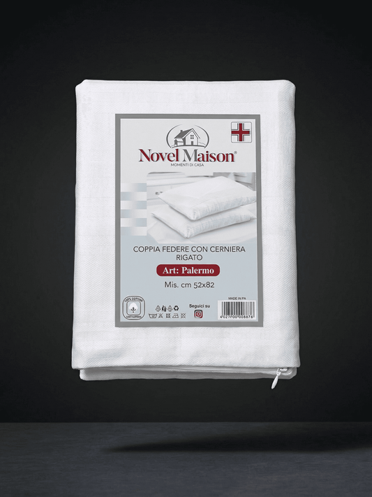 In primo piano, una fotografia di prodotto professionale e nitida del packaging di una federa per cuscino in cotone Novel Maison "copri cuscino", piegata e sigillata in un imballaggio di plastica e cartone. L'imballaggio in cartone mostra chiaramente il logo "NOVEL MAISON" con un'icona di una piccola casa e un dettaglio della bandiera italiana (o indicazione di "Made in Italy"). Il testo italiano sull'imballaggio dice "COPRI CUSCINO Con cerniera In cotone Mis. cm 50x80 1 federa". Sull'imballaggio c'è anche 