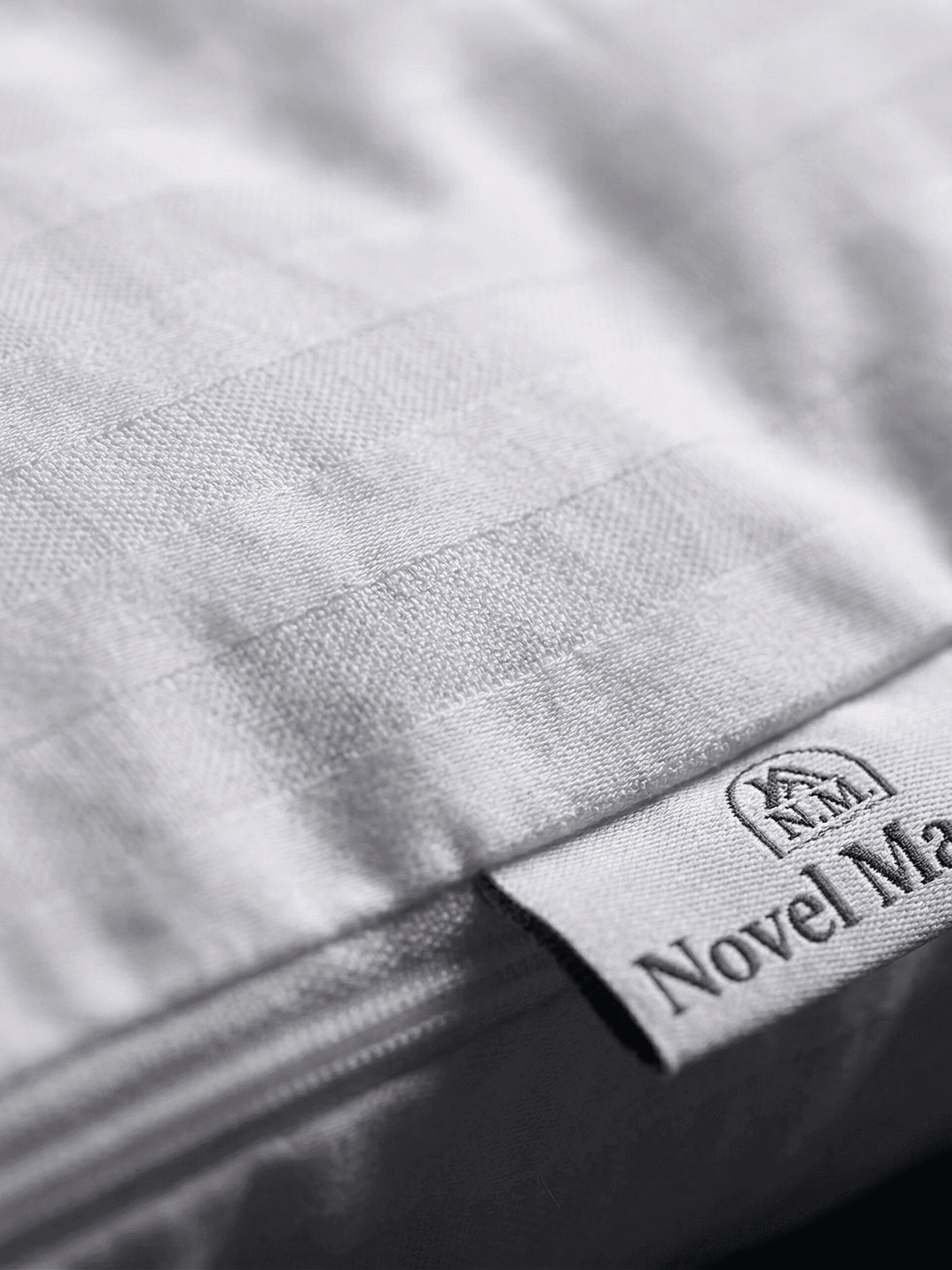 Un primo piano macro e ultra-dettagliato della trama del tessuto a coste (percale) di una federa in cotone bianco Novel Maison. L'immagine si concentra sull'orlo e sul dettaglio della cucitura. È chiaramente visibile una piccola etichetta di stoffa bianca cucita sull'orlo, che mostra il testo nero "NOVEL MAISON" e un piccolo logo a forma di casa. La cerniera invisibile è parzialmente visibile e cucita con precisione lungo il bordo. L'illuminazione è ravvicinata e soffusa, mettendo in risalto la consistenza 