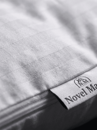Un primo piano macro e ultra-dettagliato della trama del tessuto a coste (percale) di una federa in cotone bianco Novel Maison. L'immagine si concentra sull'orlo e sul dettaglio della cucitura. È chiaramente visibile una piccola etichetta di stoffa bianca cucita sull'orlo, che mostra il testo nero "NOVEL MAISON" e un piccolo logo a forma di casa. La cerniera invisibile è parzialmente visibile e cucita con precisione lungo il bordo. L'illuminazione è ravvicinata e soffusa, mettendo in risalto la consistenza 